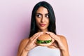 Beautiful hispanic woman eating a tasty classic burger smiling looking to the side and staring away thinking Royalty Free Stock Photo