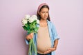 Beautiful hispanic woman expecting a baby holding flowers clueless and confused expression