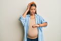 Beautiful hispanic woman expecting a baby, touching pregnant belly looking at the watch time worried, afraid of getting late