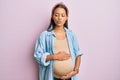 Beautiful hispanic woman expecting a baby, touching pregnant belly making fish face with mouth and squinting eyes, crazy and