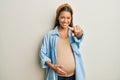 Beautiful hispanic woman expecting a baby, touching pregnant belly pointing to you and the camera with fingers, smiling positive Royalty Free Stock Photo