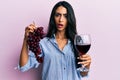 Beautiful hispanic woman holding branch of fresh grapes and red wine in shock face, looking skeptical and sarcastic, surprised Royalty Free Stock Photo