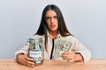 Beautiful hispanic woman holding charity jar with dollars in shock face, looking skeptical and sarcastic, surprised with open Royalty Free Stock Photo