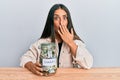 Beautiful hispanic woman holding charity jar with money covering mouth with hand, shocked and afraid for mistake Royalty Free Stock Photo