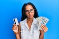 Beautiful hispanic woman holding dollars and credit card in shock face, looking skeptical and sarcastic, surprised with open mouth Royalty Free Stock Photo