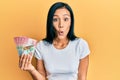 Beautiful hispanic woman holding 100 new zealand dollars banknote scared and amazed with open mouth for surprise, disbelief face Royalty Free Stock Photo