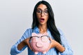 Beautiful hispanic woman holding piggy bank with glasses afraid and shocked with surprise and amazed expression, fear and excited Royalty Free Stock Photo