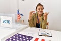 Beautiful hispanic woman at political campaign by voting ballot pointing thumb up to the side smiling happy with open mouth