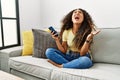 Beautiful hispanic woman sitting on the sofa at home using smartphone crazy and mad shouting and yelling with aggressive Royalty Free Stock Photo