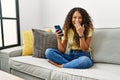 Beautiful hispanic woman sitting on the sofa at home using smartphone laughing and embarrassed giggle covering mouth with hands, Royalty Free Stock Photo