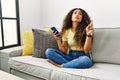 Beautiful hispanic woman sitting on the sofa at home using smartphone pointing up looking sad and upset, indicating direction with Royalty Free Stock Photo