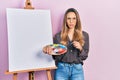 Beautiful hispanic woman standing drawing with palette by painter easel stand skeptic and nervous, frowning upset because of