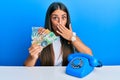 Beautiful hispanic woman with vintage telephone holding australian dollars covering mouth with hand, shocked and afraid for Royalty Free Stock Photo