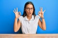 Beautiful hispanic woman wearing casual clothes sitting on the table looking surprised and shocked doing ok approval symbol with Royalty Free Stock Photo