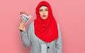 Beautiful hispanic woman wearing islamic hijab holding australian dollars banknotes thinking attitude and sober expression looking