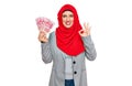 Beautiful hispanic woman wearing islamic hijab holding chinese yuan banknotes doing ok sign with fingers, smiling friendly