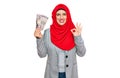 Beautiful hispanic woman wearing islamic hijab holding japanese yen banknotes doing ok sign with fingers, smiling friendly