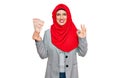 Beautiful hispanic woman wearing islamic hijab holding turkish lira banknotes doing ok sign with fingers, smiling friendly