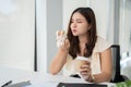 A beautiful, hungry Asian businesswoman is eating a sandwich and drinking coffee at her desk