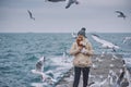 Beautiful image of young smiling woman feeds seagulls on the sea. Pretty female wearing coat, scarf, hat watching flying seagulls Royalty Free Stock Photo