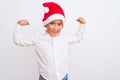Beautiful kid boy wearing Christmas Santa hat standing over isolated white background showing arms muscles smiling proud Royalty Free Stock Photo