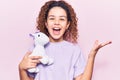 Beautiful kid girl with curly hair holding animal doll toy celebrating victory with happy smile and winner expression with raised
