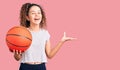 Beautiful kid girl with curly hair holding basketball ball celebrating victory with happy smile and winner expression with raised