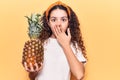 Beautiful kid girl with curly hair holding pineapple covering mouth with hand, shocked and afraid for mistake Royalty Free Stock Photo