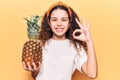 Beautiful kid girl with curly hair holding pineapple doing ok sign with fingers, smiling friendly gesturing excellent symbol Royalty Free Stock Photo