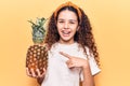 Beautiful kid girl with curly hair holding pineapple smiling happy pointing with hand and finger Royalty Free Stock Photo