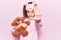 Beautiful kid girl with curly hair wearing sleep mask and pajamas holding teddy bear with open hand doing stop sign with serious