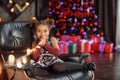 Beautiful kid girl 5-6 year old wearing stylish dress sitting in armchair over Christmas tree in room. Looking at camera. Holiday