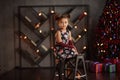 Beautiful kid girl 5-6 year old wearing stylish dress sitting in armchair over Christmas tree in room. Looking at camera. Holiday