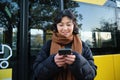 Beautiful korean girl, student on bus stop, looking at her smartphone, checking timetable, reading text message, wearing Royalty Free Stock Photo