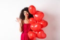 Beautiful lady with curly hair, standing near valentines holiday balloons and waving hand, saying hello, standing