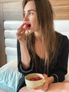 Beautiful lady in hotel bed eats strawberry in black pajamas and looking away