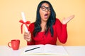 Beautiful latin young woman with long hair holding graduate degree diploma scared and amazed with open mouth for surprise, Royalty Free Stock Photo