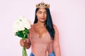 Beautiful latin young woman with long hair wearing princess crown and holding flowers thinking attitude and sober expression