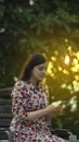 Beautiful latin young woman in white floral design dress reading on her smartphone in the garden at sunset with the sun`s rays Royalty Free Stock Photo