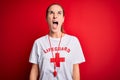 Beautiful lifeguard woman wearing t-shirt with red cross using whistle over isolated background angry and mad screaming frustrated