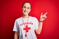 Beautiful lifeguard woman wearing t-shirt with red cross using whistle over isolated background cheerful with a smile on face