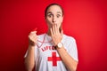 Beautiful lifeguard woman wearing t-shirt with red cross using whistle over isolated background cover mouth with hand shocked with