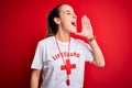 Beautiful lifeguard woman wearing t-shirt with red cross using whistle over isolated background shouting and screaming loud to