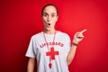 Beautiful lifeguard woman wearing t-shirt with red cross using whistle over isolated background Surprised pointing with finger to