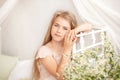 Beautiful little blonde girl with long hair sits on a bed with a bouquet of daisies in a bright white bedroom with a rustic interi