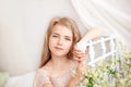 Beautiful little blonde girl with long hair sits on a bed with a bouquet of daisies in a bright white bedroom with a rustic interi Royalty Free Stock Photo