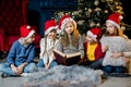 Beautiful little children read a book with Christmas stories on the background of a Christmas tree in red New Year hats