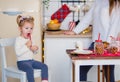 A beautiful little daughter with blond hair in a white sweater sits in the kitchen, eats a red caramel cane. christmas home decor