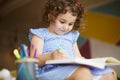 Beautiful little girl with dark curly hair in dress dreamily drawing in coloring book with felt-tip pens on sofa at home