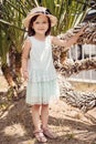 Beautiful little girl in a hat and a white dress stands near a palm tree. Royalty Free Stock Photo